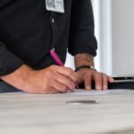 Homme écrivant sur une table avec un stylo, dossier et porte visibles