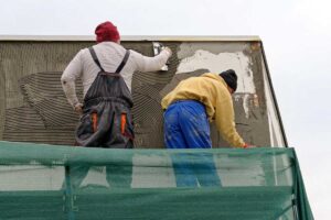 Deux ouvriers appliquent de l'enduit blanc sur la façade d'un bâtiment avec des outils professionnels