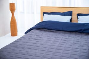 Lit avec draps bleus, oreillers, tablette en bois, lampe, chambre claire