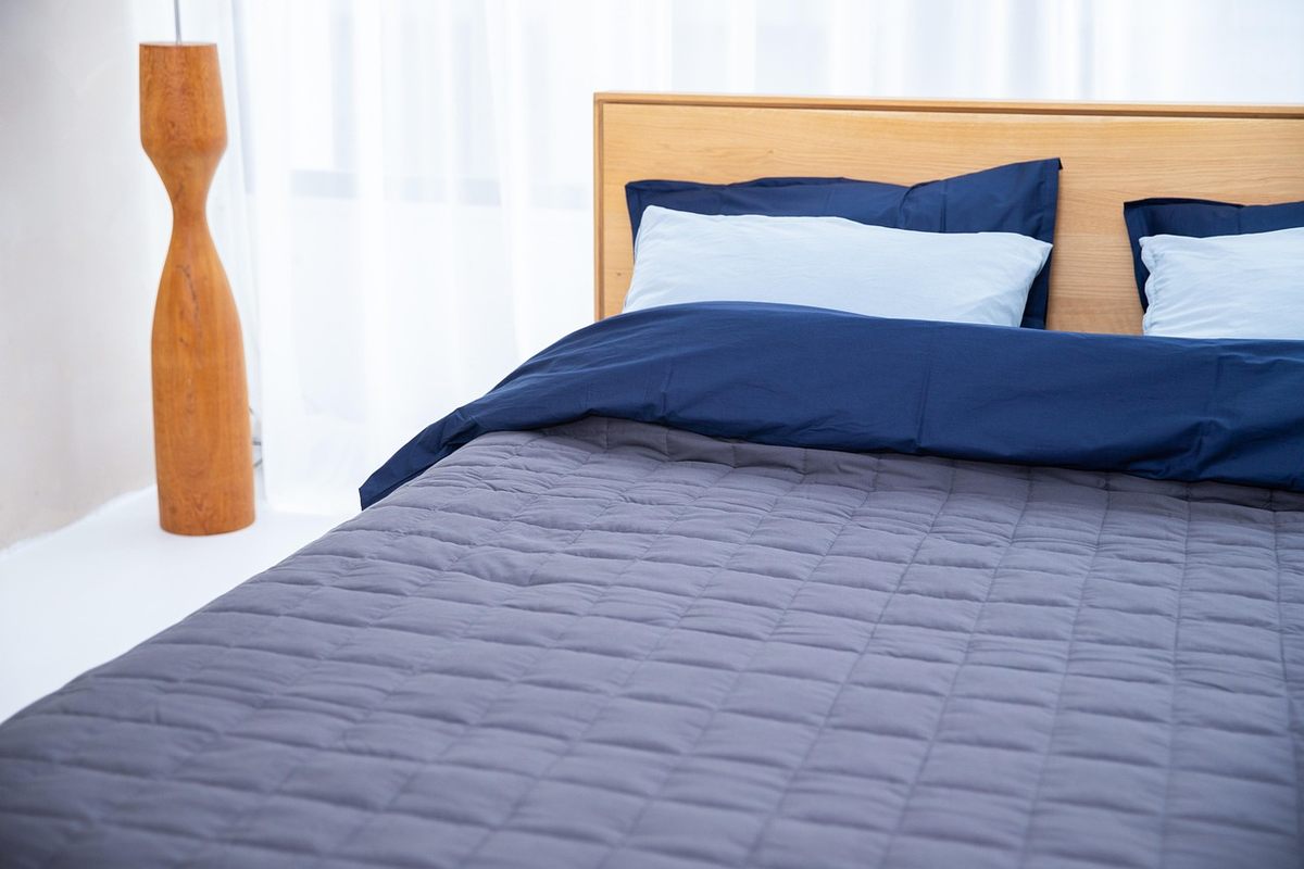 Lit avec draps bleus, oreillers, tablette en bois, lampe, chambre claire